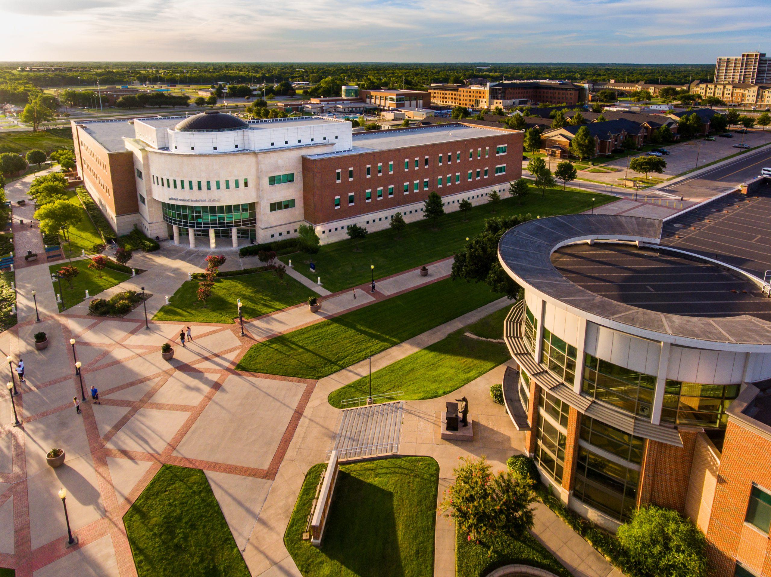 Aerial view of the East Texas A&M University campus, showcasing modern buildings, walkways, and green spaces under a clear sky.